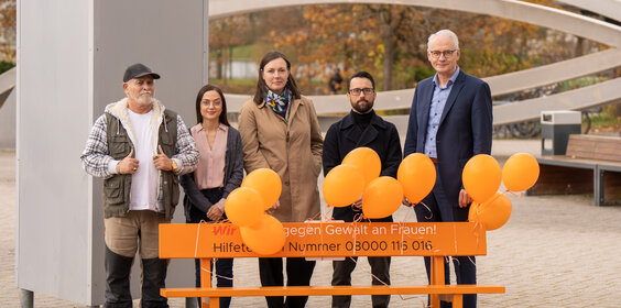 Fünf Personen stehen hinter einer orangen Bank. Daran sind orange Luftballons befestigt und auf der Bank ein Aufdruck mit der Notruf-Telefonnummer 08000116016 für Gewalt gegen Frauen.