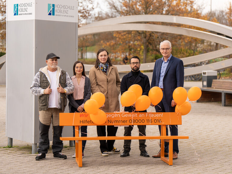 Fünf Personen stehen hinter einer orangen Bank. Daran sind orange Luftballons befestigt und auf der Bank ein Aufdruck mit der Notruf-Telefonnummer 08000116016 für Gewalt gegen Frauen.