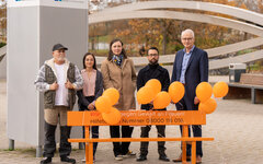 Fünf Personen stehen hinter einer orangen Bank. Daran sind orange Luftballons befestigt und auf der Bank ein Aufdruck mit der Notruf-Telefonnummer 08000116016 für Gewalt gegen Frauen.