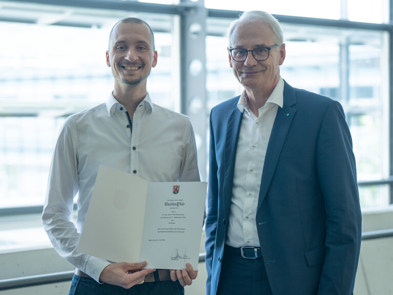 Zwei Herren stehen beieinander. Der Linke von Beiden hält eine Urkunde in der Hand.