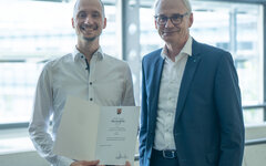 Zwei Herren stehen beieinander. Der Linke von Beiden hält eine Urkunde in der Hand.