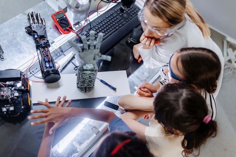 Drei Mädchen und eine Frau sitzen an einem Experimentiertisch. Darauf stehen Roboterhände.