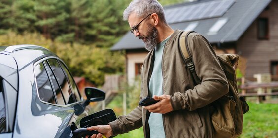 EIn Mann steht mit Handy und Ladekabel an seinem Elektroauto neben einem Haus.