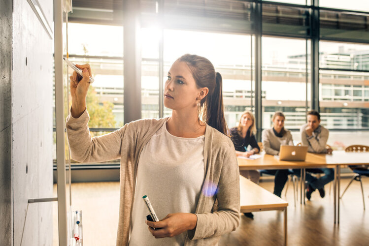 Studentin steht mit einem Stift in der Hand vor einem Whiteboard. Im Hintergrund erkennt man mehrer Studierende im Vorlesungsraum